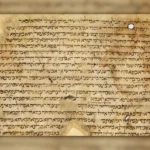 A widescreen archival-style image featuring an ancient manuscript page centered against a softly blurred background created from an enlarged version of the same manuscript. The central parchment sheet has a warm beige and golden-brown tone, with visible signs of age including faded areas, stains, worn edges, tears, holes, and irregular parchment texture. Dense handwritten text in dark brown ink fills the page, written in an ancient Hebrew or Aramaic script with closely spaced horizontal lines and variations in ink intensity caused by aging and manuscript wear. The surrounding left and right areas form a blurred pillarbox effect, extending the parchment colors and script patterns into a soft, out-of-focus backdrop that matches the central document. The main manuscript appears slightly raised from the background with a subtle shadow, creating a layered presentation that emphasizes the historical artifact. The overall composition evokes a museum display or scholarly archive, highlighting the texture, craftsmanship, and historical character of an ancient Jewish manuscript fragment.