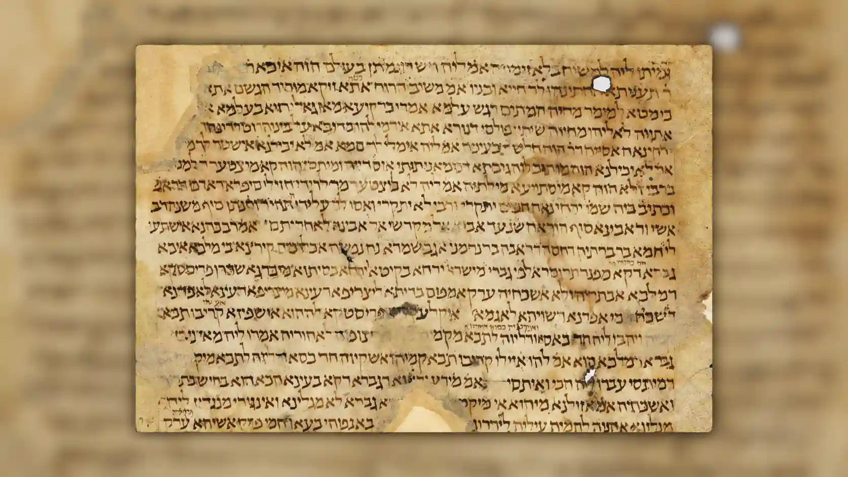 A widescreen archival-style image featuring an ancient manuscript page centered against a softly blurred background created from an enlarged version of the same manuscript. The central parchment sheet has a warm beige and golden-brown tone, with visible signs of age including faded areas, stains, worn edges, tears, holes, and irregular parchment texture. Dense handwritten text in dark brown ink fills the page, written in an ancient Hebrew or Aramaic script with closely spaced horizontal lines and variations in ink intensity caused by aging and manuscript wear. The surrounding left and right areas form a blurred pillarbox effect, extending the parchment colors and script patterns into a soft, out-of-focus backdrop that matches the central document. The main manuscript appears slightly raised from the background with a subtle shadow, creating a layered presentation that emphasizes the historical artifact. The overall composition evokes a museum display or scholarly archive, highlighting the texture, craftsmanship, and historical character of an ancient Jewish manuscript fragment.